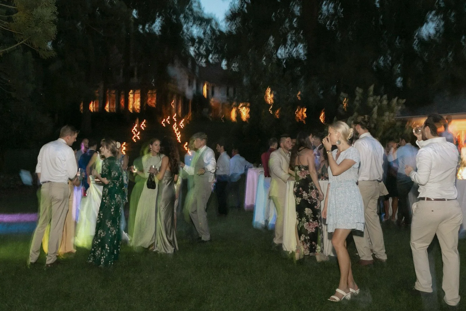 People attending an outdoor evening celebration or party, dressed in formal and semi-formal attire, with some holding drinks, under trees with lights and a fence in the background.