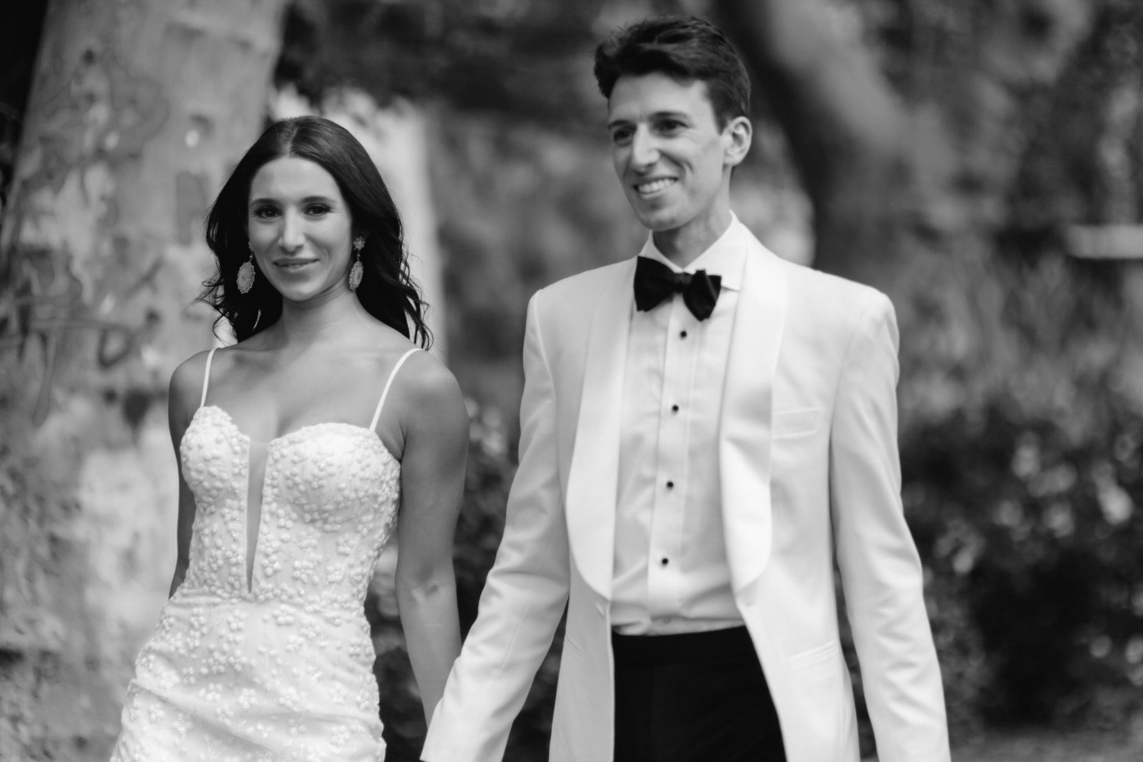 Black and white photo of a bride and groom holding hands outdoors, dressed in wedding attire, with trees in the background.