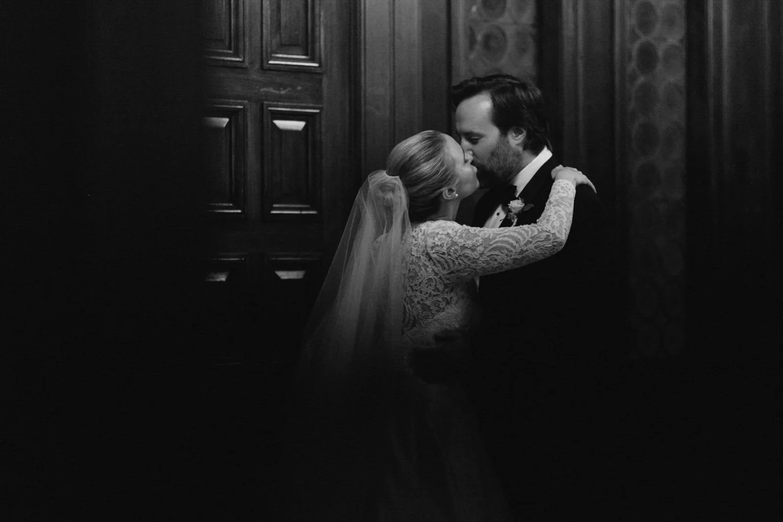 Black and white photo of a bride and groom sharing a kiss inside a dimly lit room with wooden paneling.