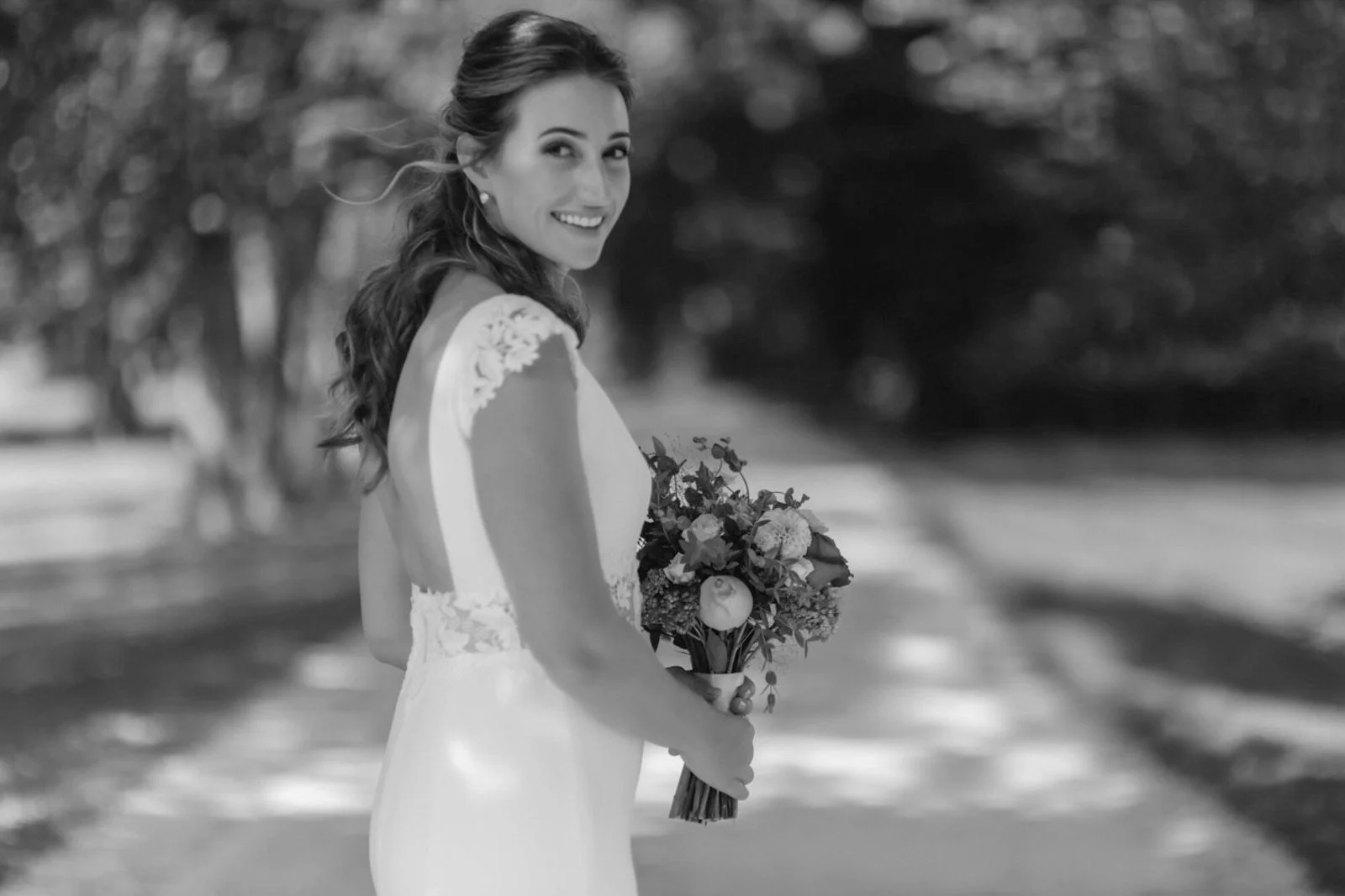 A smiling bride in a wedding dress holding a bouquet of flowers, standing outdoors on a sunny day with trees in the background.