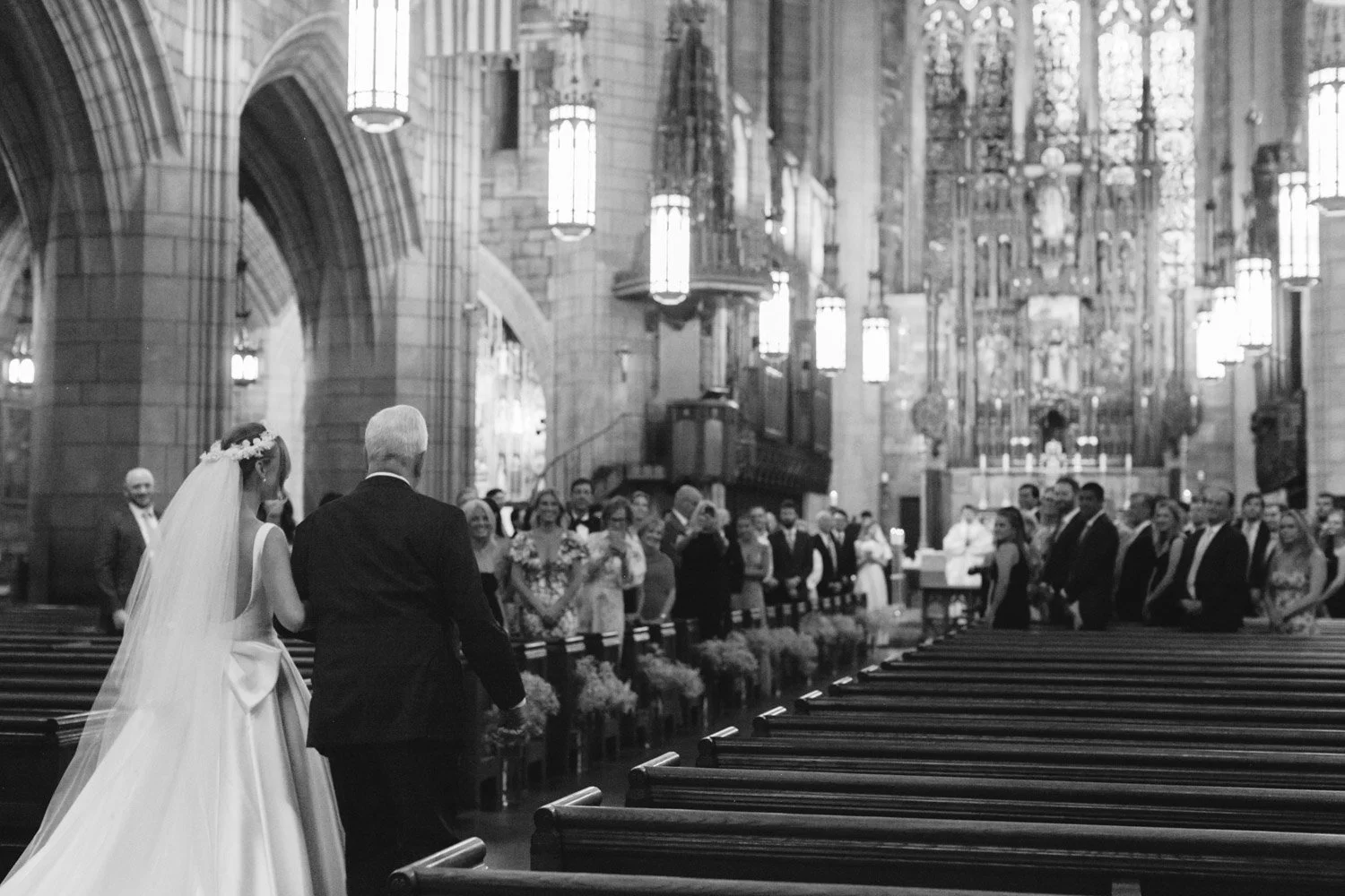 A bride and an older man walking down the aisle in a church with a large congregation watching. The church has high arched ceilings, stained glass windows, and ornate decorations at the altar.