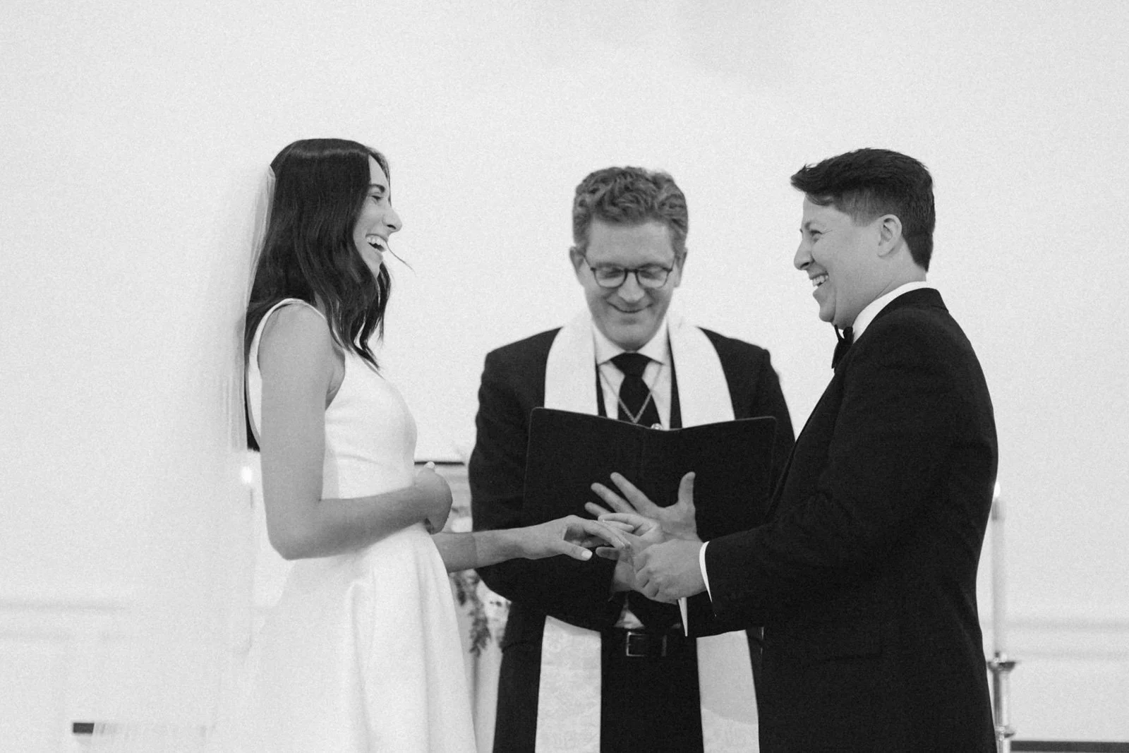 A couple getting married, the bride in a sleeveless white dress, the groom in a tuxedo, exchanging rings indoors, officiant in the middle holding a booklet, all smiling and laughing.