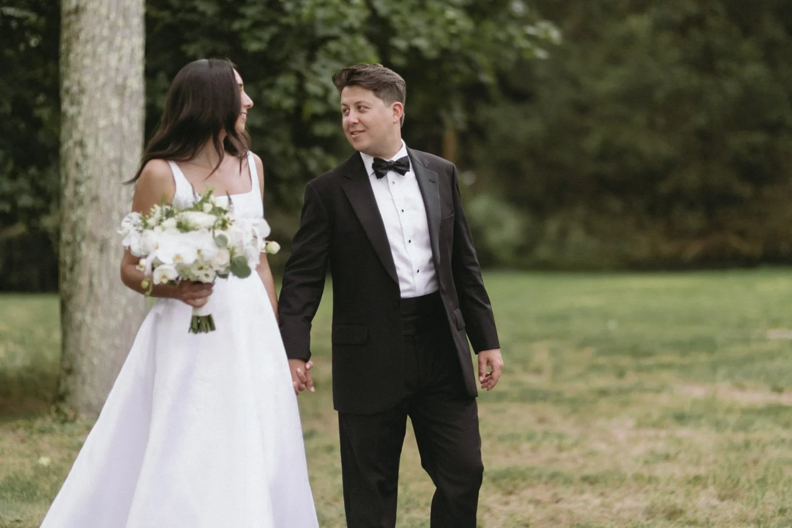 A bride and groom holding hands outdoors in a park. The bride wears a white wedding dress and holds a bouquet of white flowers, the groom wears a black tuxedo with a bow tie. They are smiling at each other.