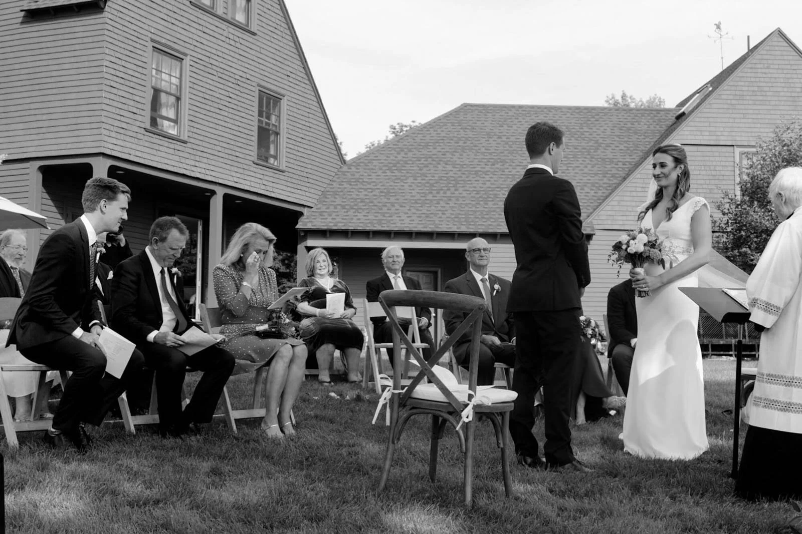 A black and white photo of a wedding ceremony outdoors, with a couple standing in front of an officiant, and friends and family seated behind them on the grass.