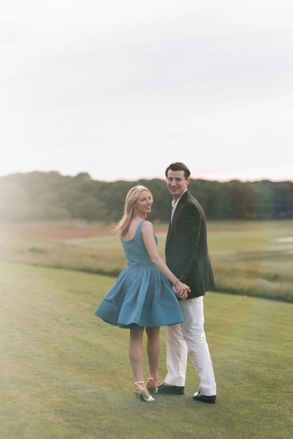 A couple standing hand in hand at The Shinnecock Golf Club. During their rehearsal dinner, smiling at the camera. The woman is in a blue dress and high heels, the man in a dark blazer and white pants.