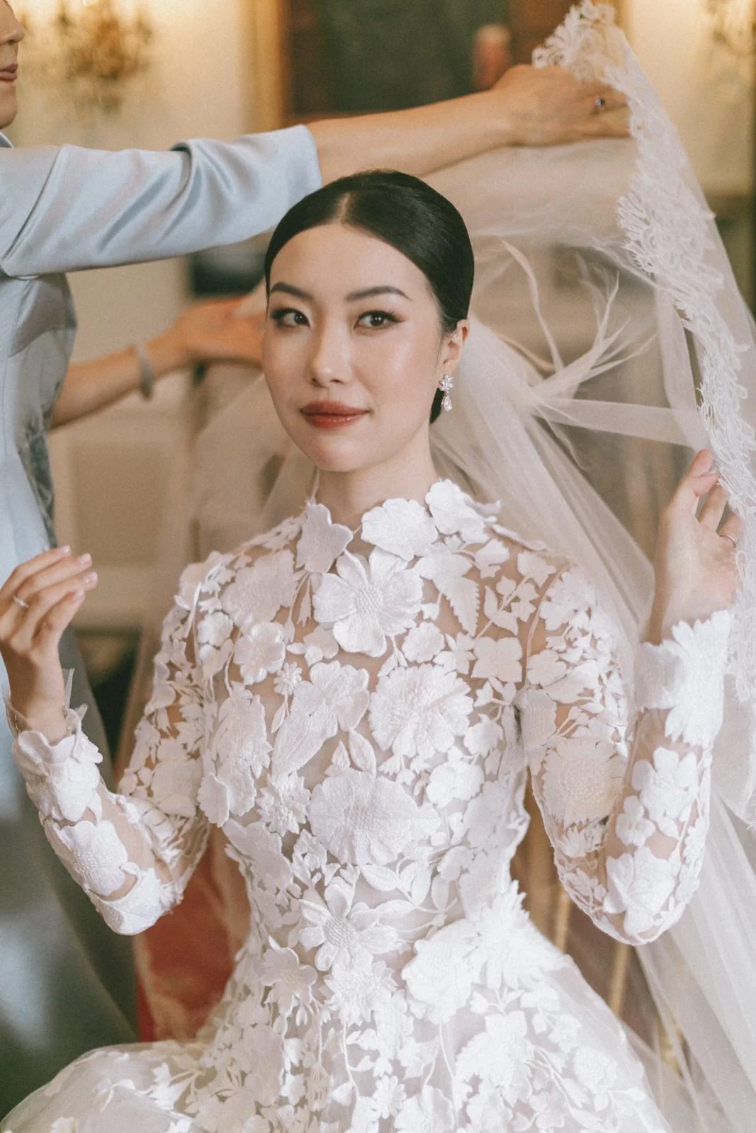 A bride in a white floral lace wedding dress with long sleeves, sitting as a stylist adjusts her veil.