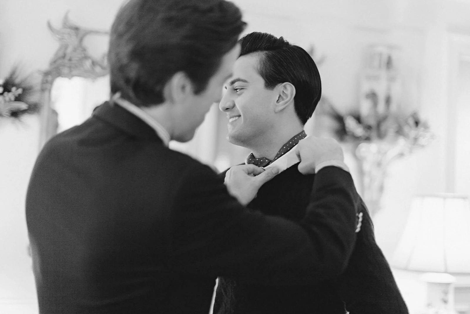 Two men dressed in tuxedos are dancing closely, smiling at each other, in a living room decorated with flowers and a lamp.
