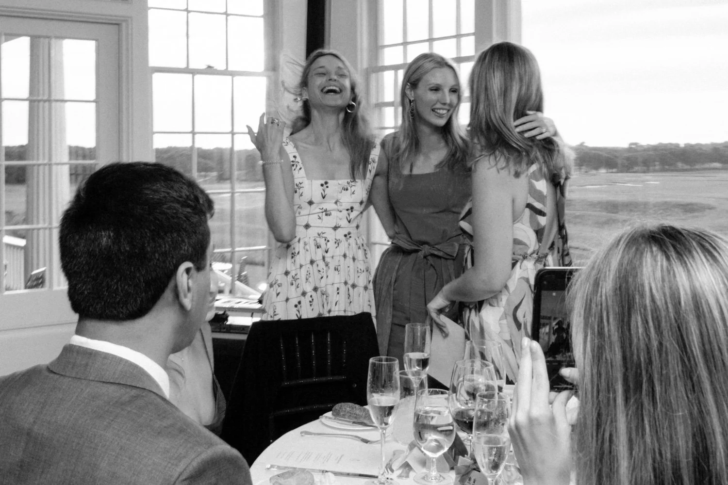 Four women two of whom are standing and two sitting at a dining table, with large windows and scenic view in the background, in a black and white photo, appearing to be at a celebratory event.