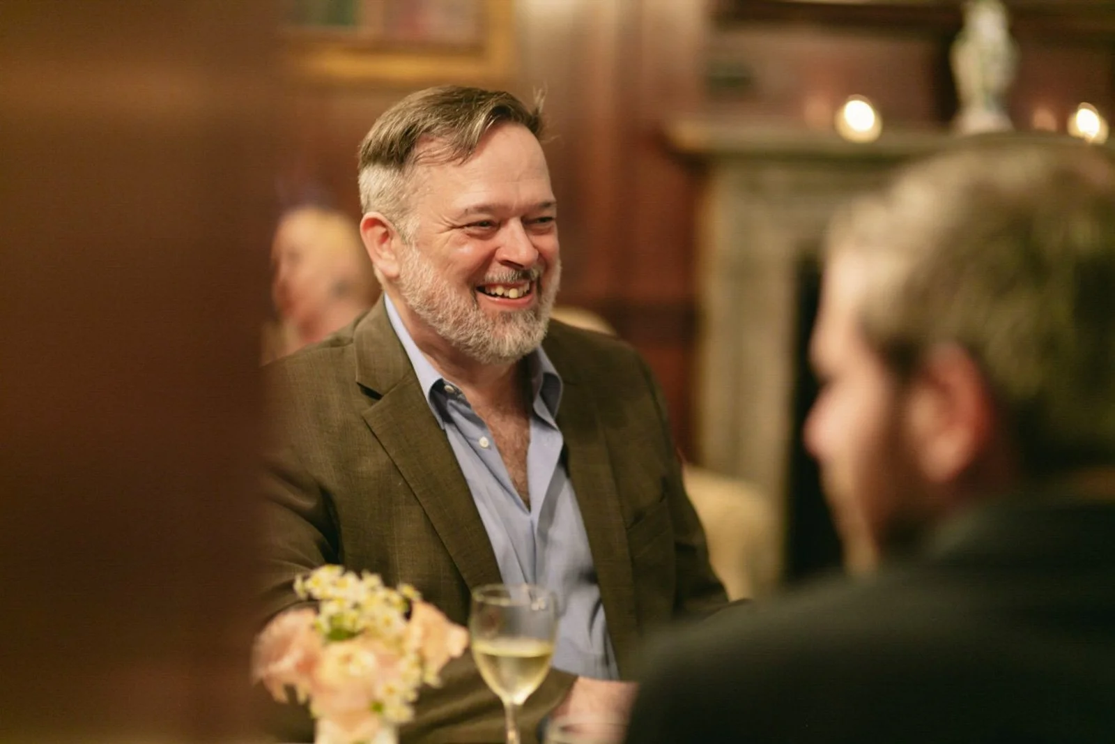 A smiling man with a beard and gray hair, wearing a brown blazer and light blue shirt, engaging in conversation at a social gathering in a warmly lit room, with a woman in the foreground and a glass of white wine on the table.