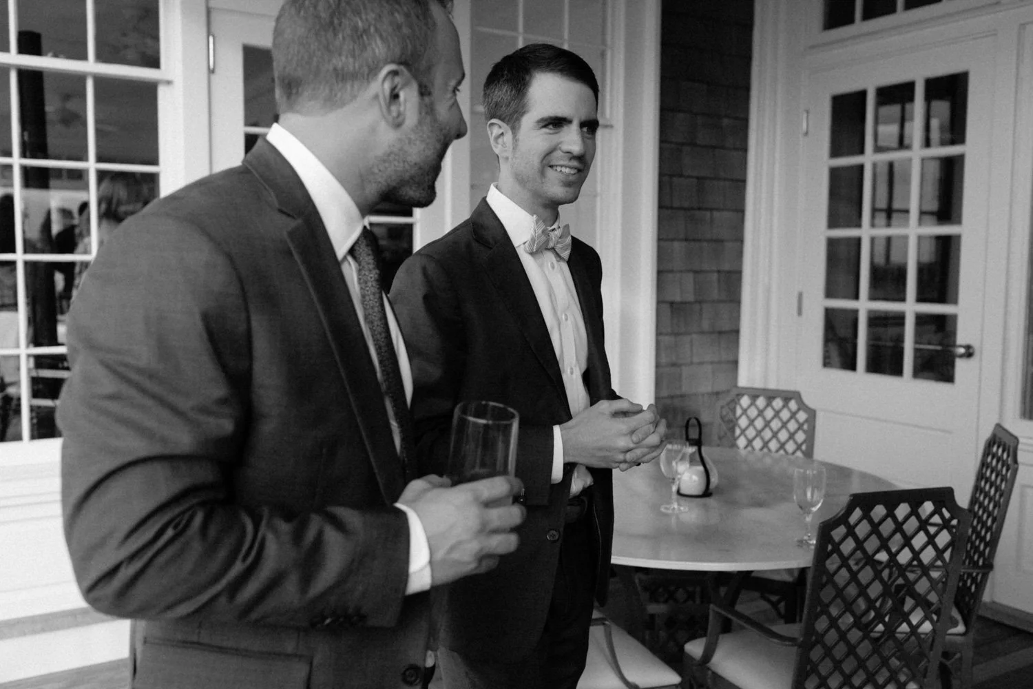 Two men in suits and bow ties talking in a dining room, one holding a glass of wine.