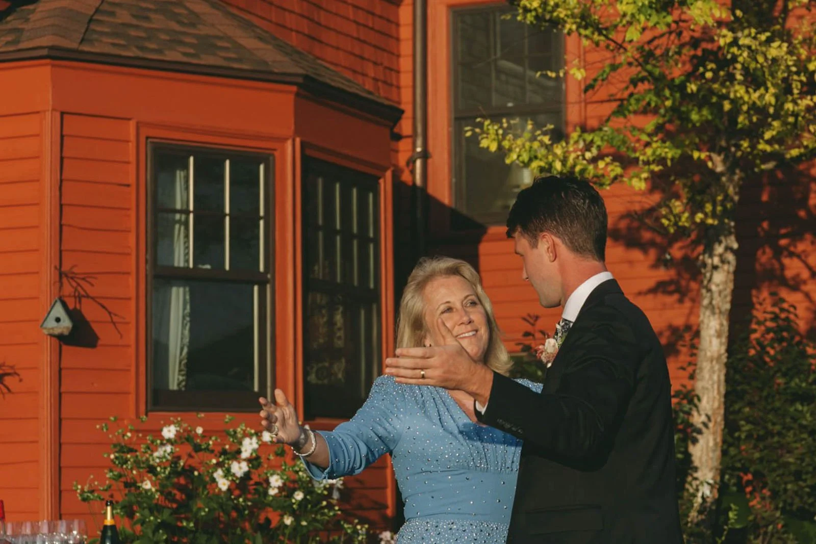 A woman in a sparkly blue dress dancing with a man in a black tuxedo outdoors in front of a red house, during a celebration at sunset.