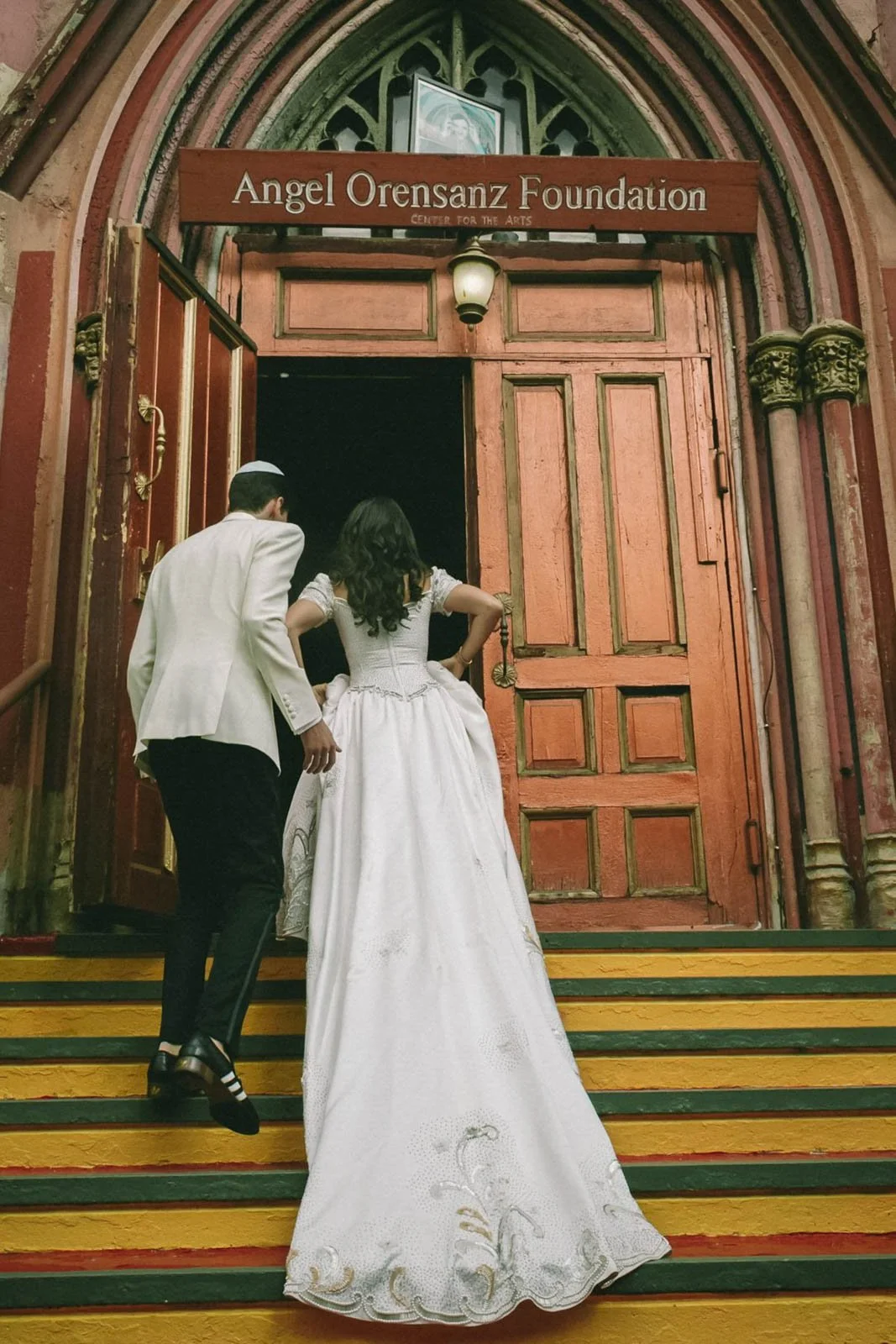 People dressed in traditional Jewish wedding attire ascending the steps of a historical church with a wooden sign that reads 'Angel Orensanz Foundation, Center for the Arts.'