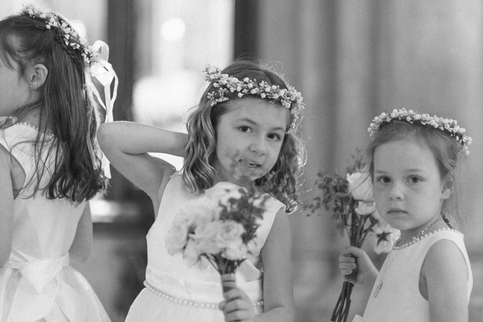 Black and white photo of three young girls dressed in formal attire, each wearing a floral crown and holding flower bouquets, possibly at a wedding or special event.