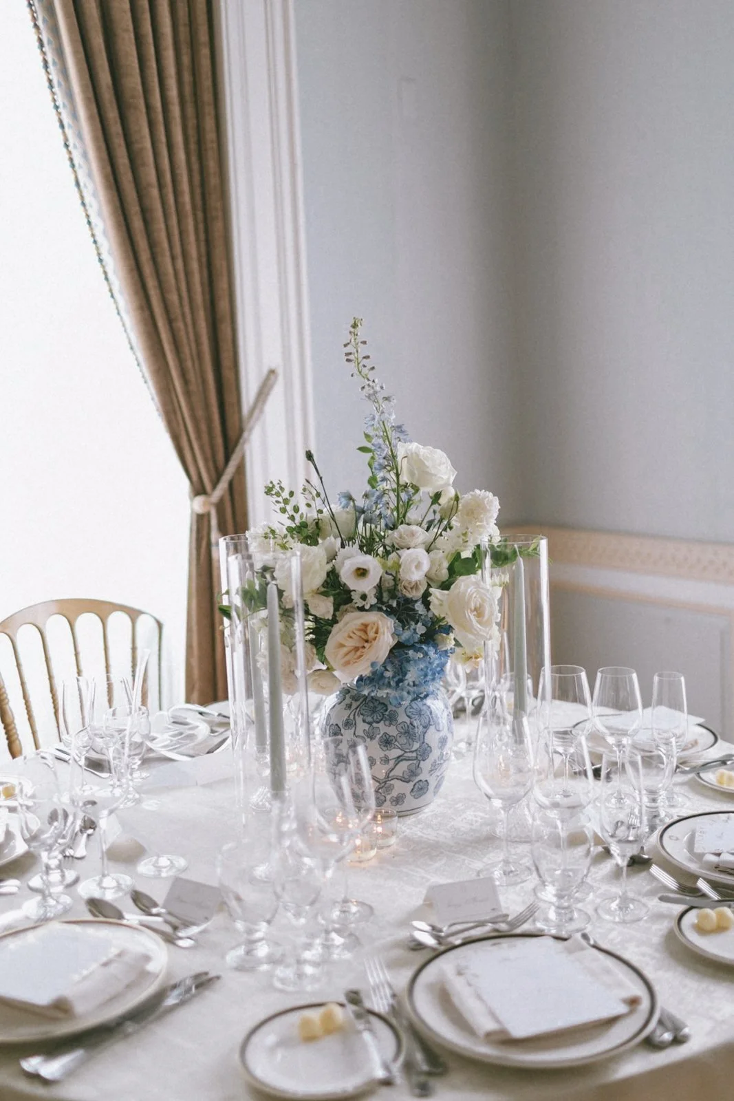 A formal dining table set for a special occasion, with a white tablecloth, elegant china, crystal glasses, and a floral centerpiece with white and blue flowers in a decorative white vase.