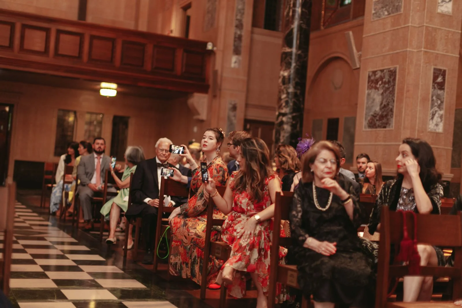 A group of people dressed in elegant attire sitting on chairs inside a decorated hall. Some are taking photos while others are engaged in conversations, with a checkered black and white floor and ornate woodwork and marble columns in the background.