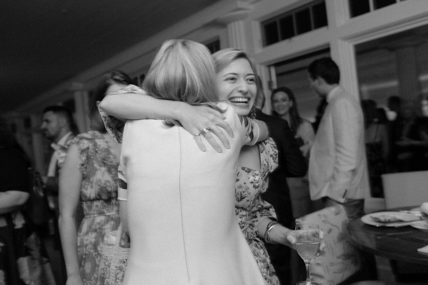 Two women hugging at a social gathering, with one woman smiling and holding a glass of wine, in an indoor setting with other people in the background.