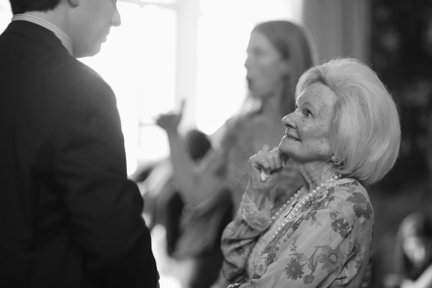 An elderly woman with white hair and a floral blouse smiling and touching her chin, engaged in conversation with a man in a suit; two women are in the background, one gesturing with her hand.
