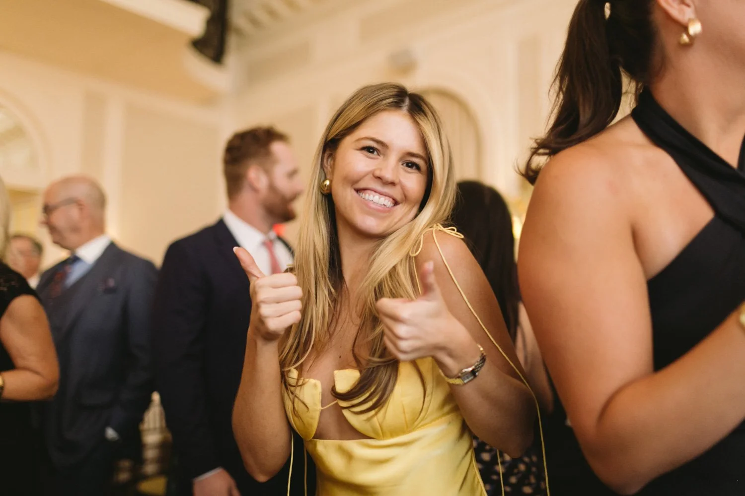 A smiling woman in a yellow dress giving a thumbs-up at a social event or party, surrounded by other people dressed in formal attire.