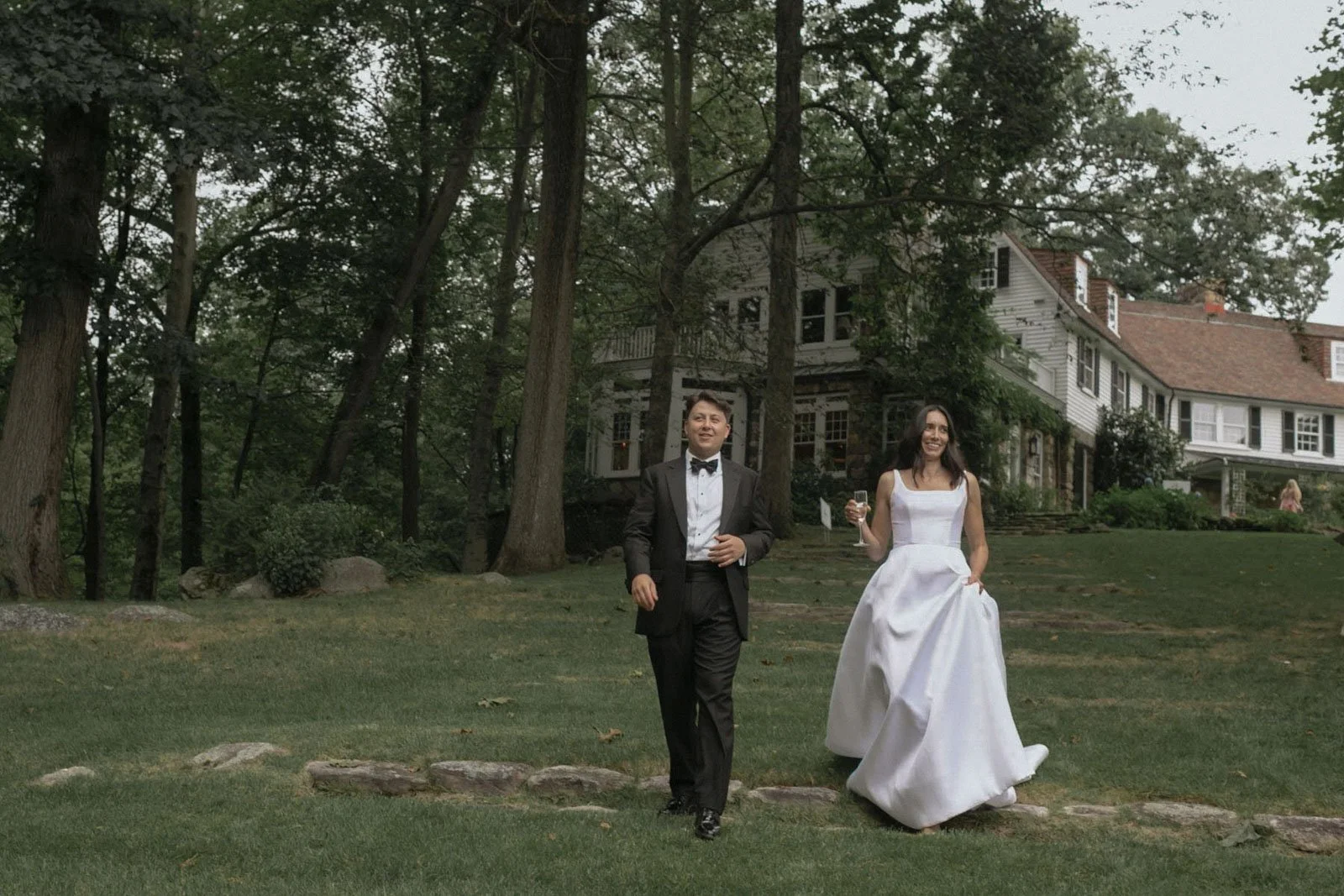 A man in a tuxedo and a woman in a wedding dress walking across a lawn holding a glass of champagne, with a large house and trees in the background.