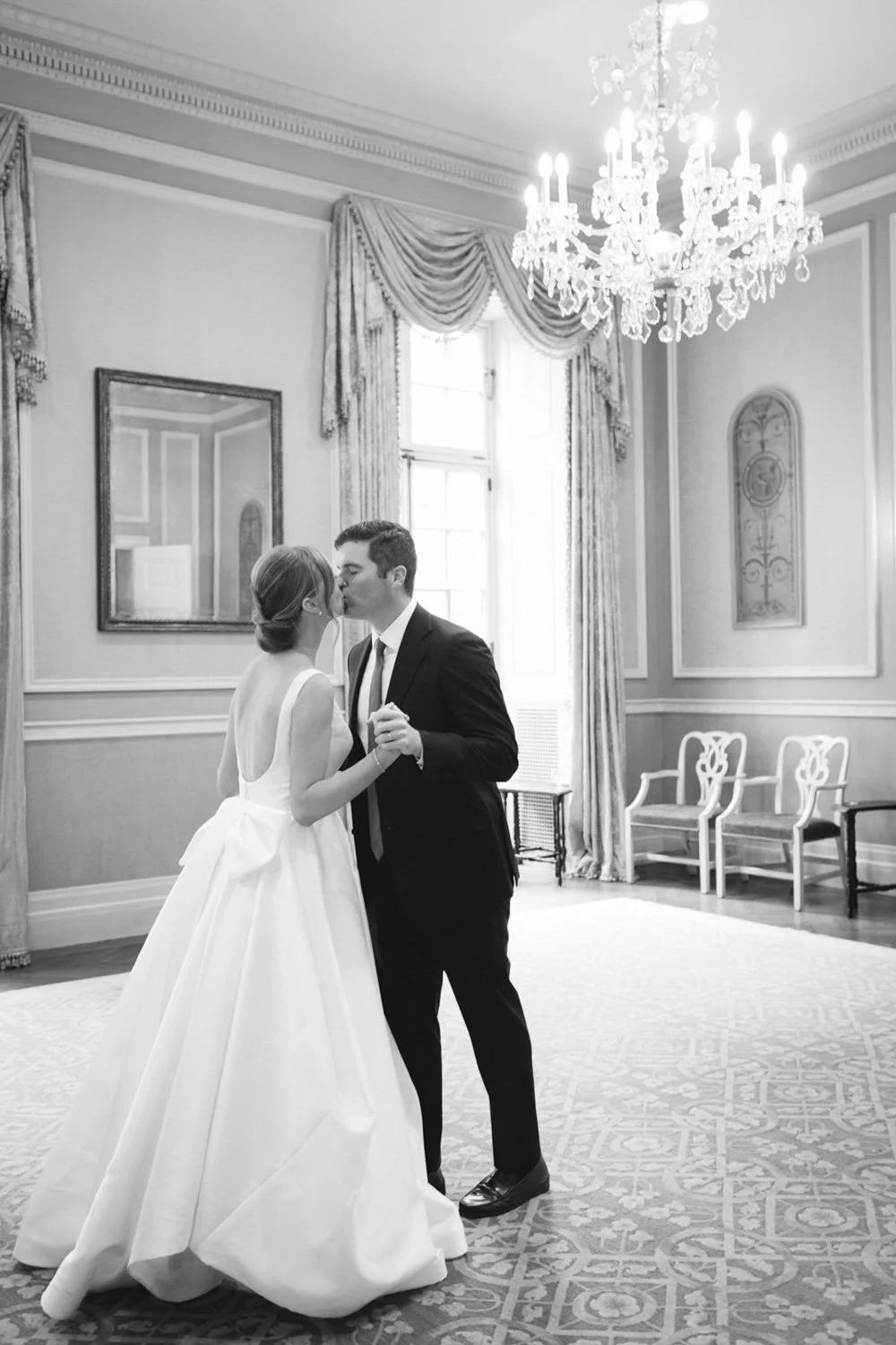 A bride and groom sharing a kiss and holding hands inside a formal, elegant room with a chandelier, large windows, and ornate curtains.