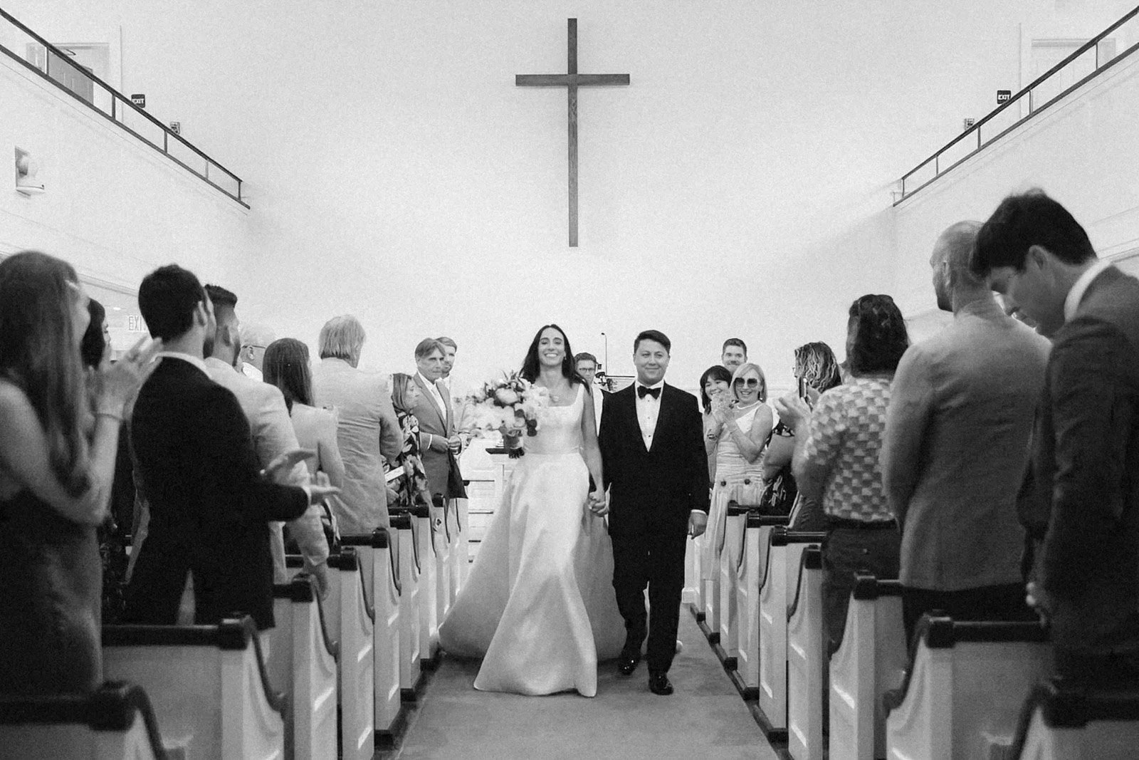 A bride and groom walking down the aisle in a church with guests on either side. The bride is holding a bouquet, and a cross is hanging on the wall behind them.