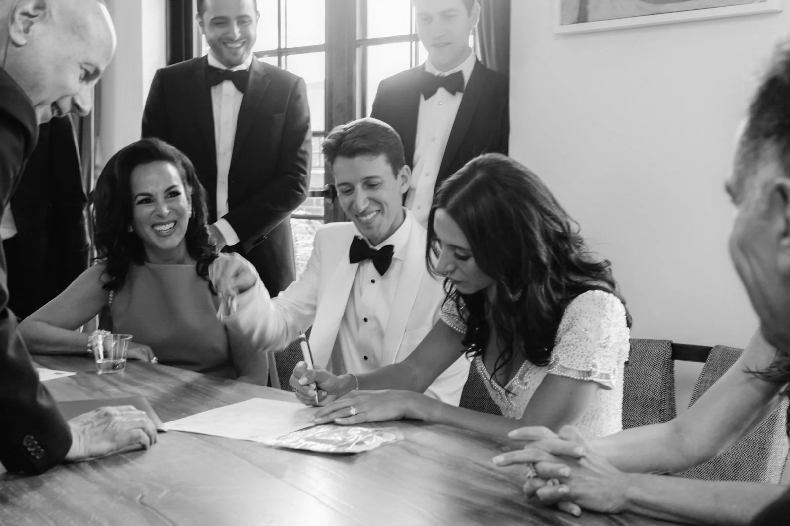 A wedding ceremony with a bride and groom signing a marriage document surrounded by friends and family, all dressed in formal attire.