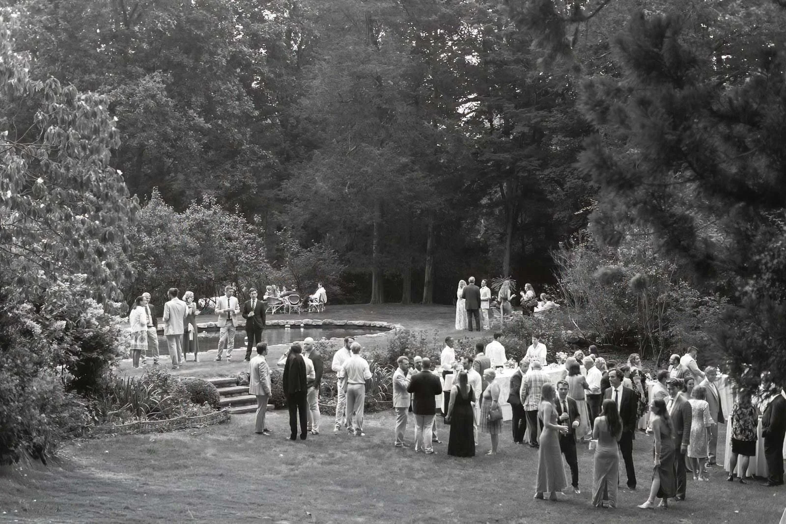 Black and white photo of a large outdoor gathering of people in a garden, with some near a small pond, some standing and talking, and others sitting at tables, surrounded by trees and bushes.