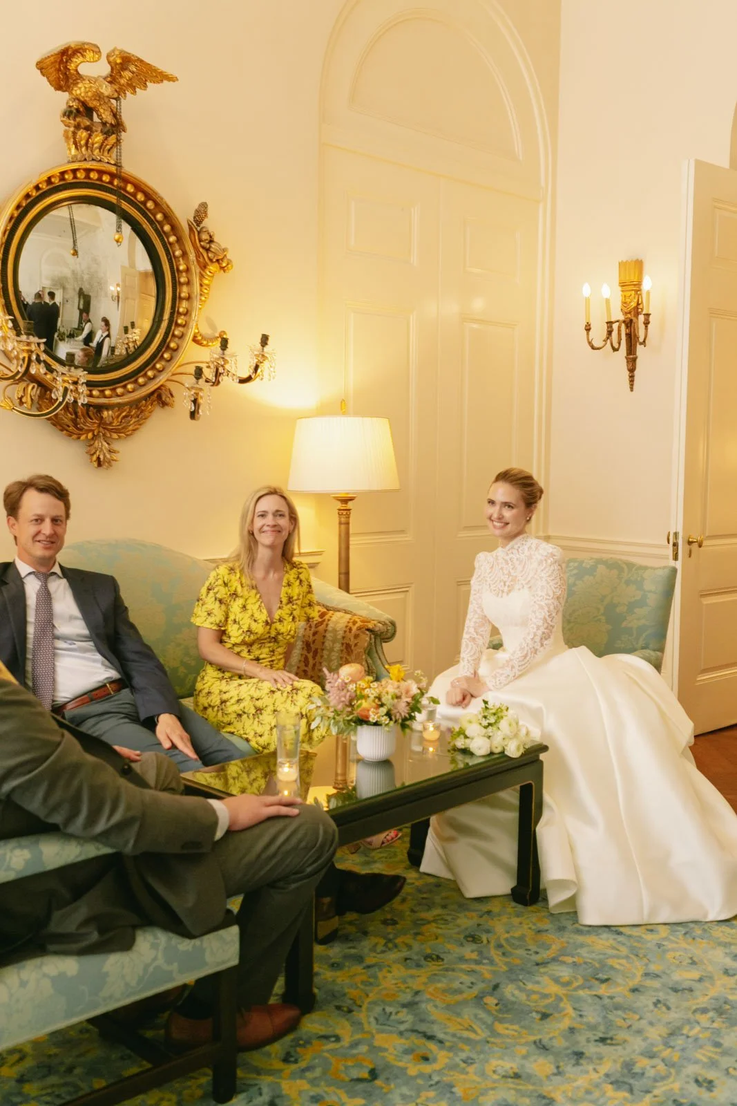A bride in a white wedding dress sitting on a sofa with three guests in a decorated, elegant room with a mirror, chandelier, and floral arrangements.