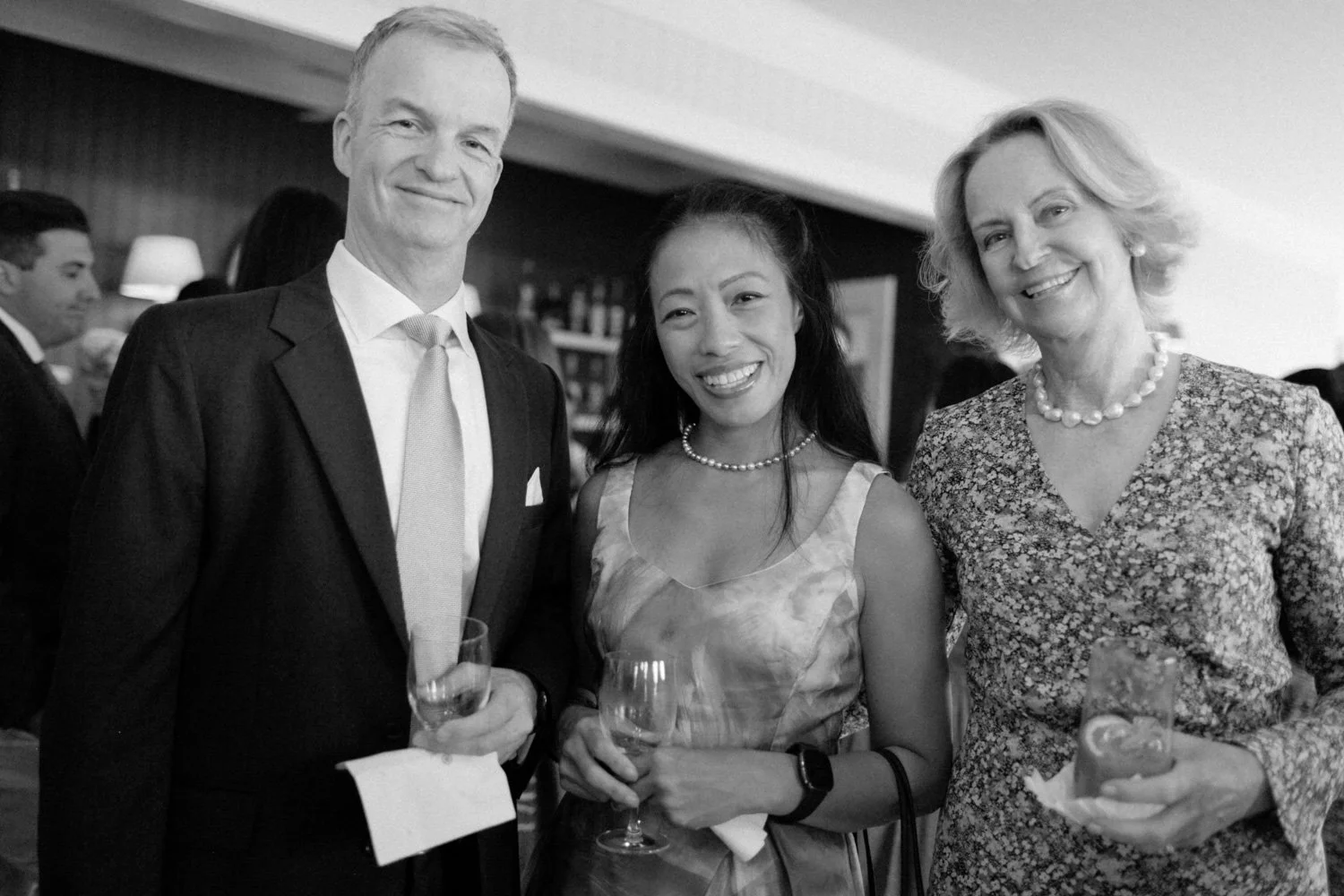 Three people, two women and one man, at a social gathering, dressed in formal attire, holding glasses of wine or cocktails, smiling for the camera in black and white.