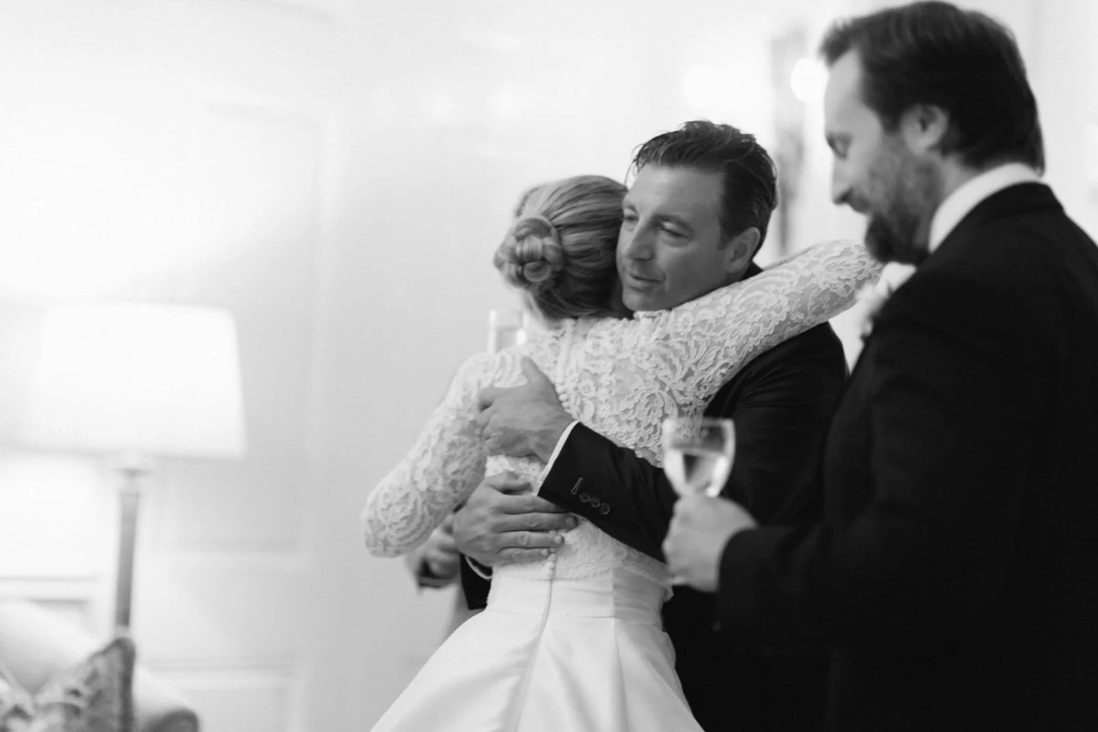A black and white photo of a bride hugging a groom at a wedding reception, with a man in a suit holding a champagne flute nearby.