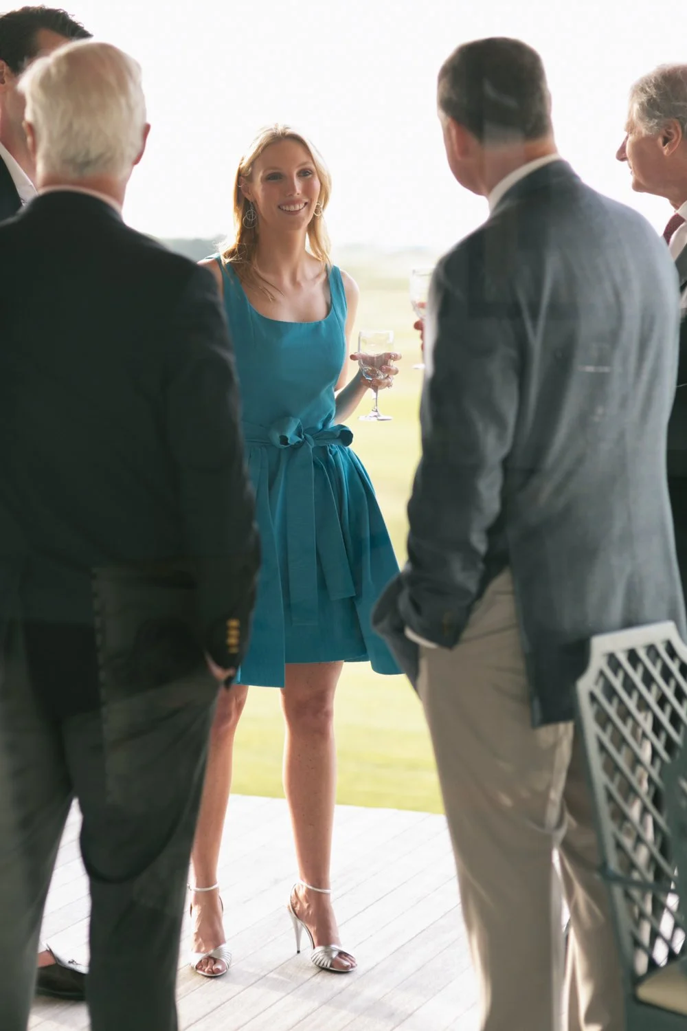 A woman in a blue dress holding a glass of white wine, smiling and talking with a group of men in suits at an outdoor event.
