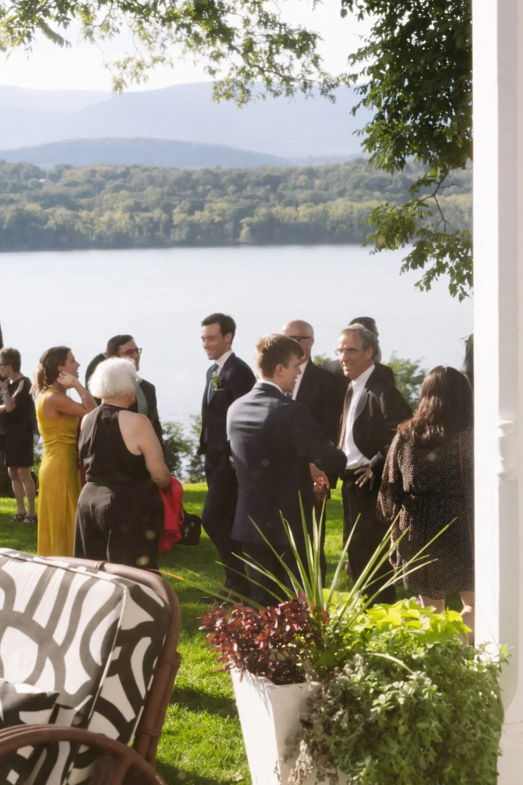 A group of people dressed in formal attire socializing outdoors near a large body of water with trees and mountains in the background. There are potted plants and a patterned chair in the foreground.