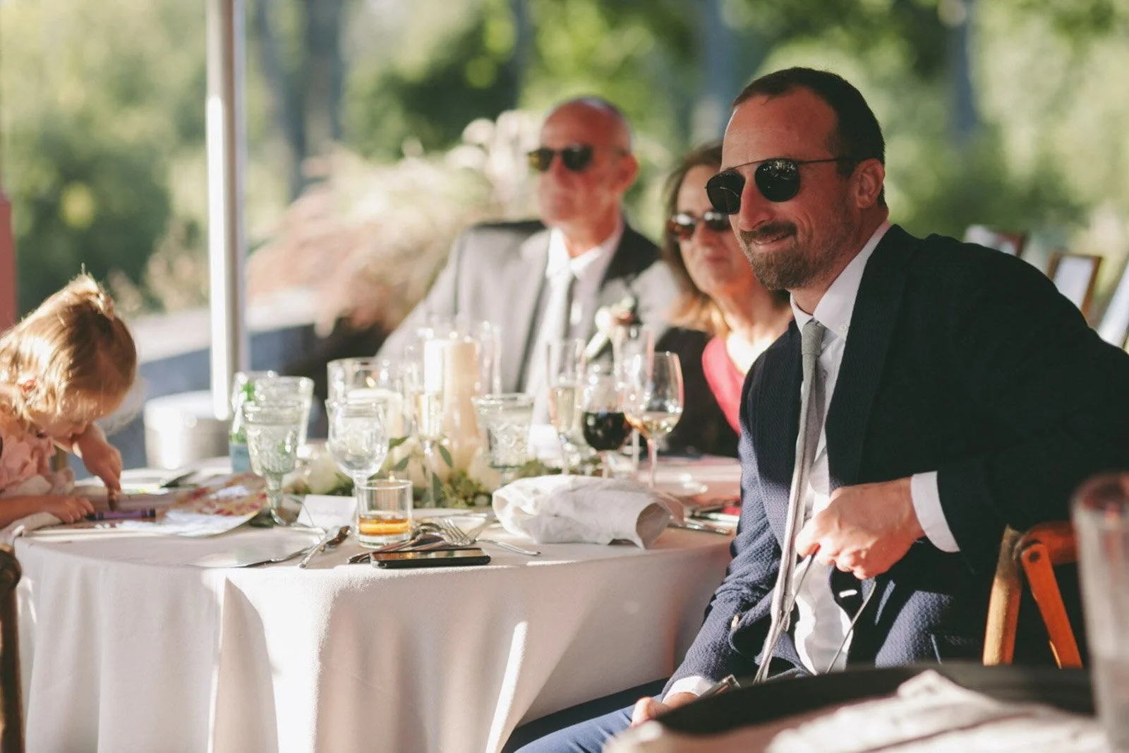 A group of people dressed in formal attire, sitting at a banquet table outdoors during daytime, with glasses, candles, and decorations on the table.
