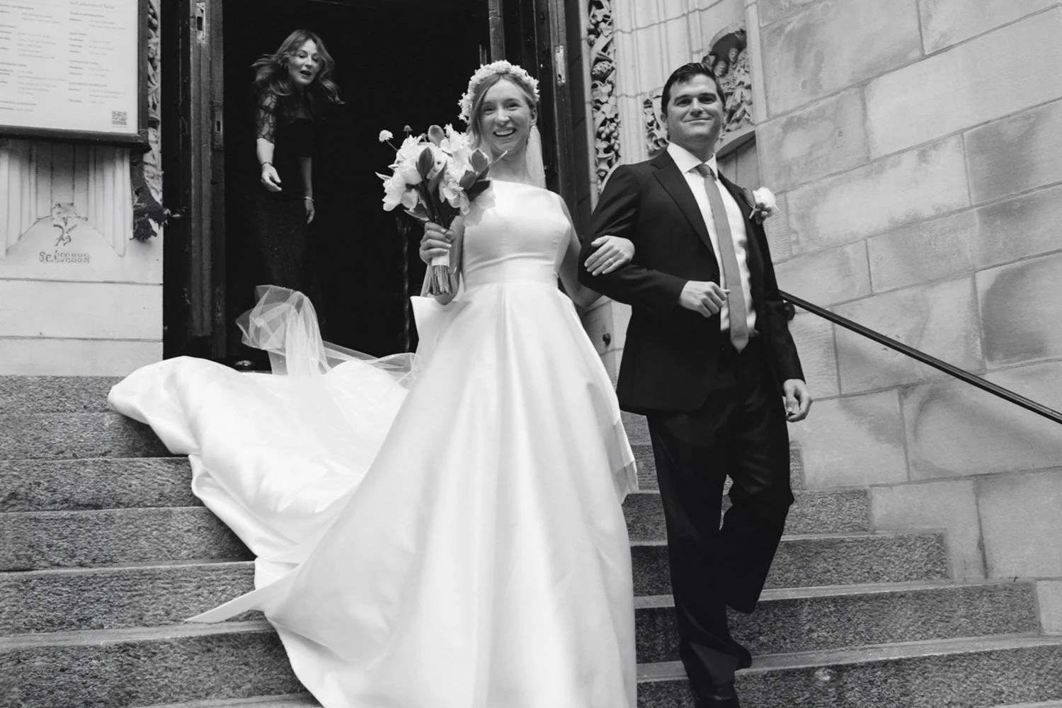 Black and white photo of a bride and groom descending the steps of a church after their wedding, with the bride holding a bouquet of flowers and the groom smiling.