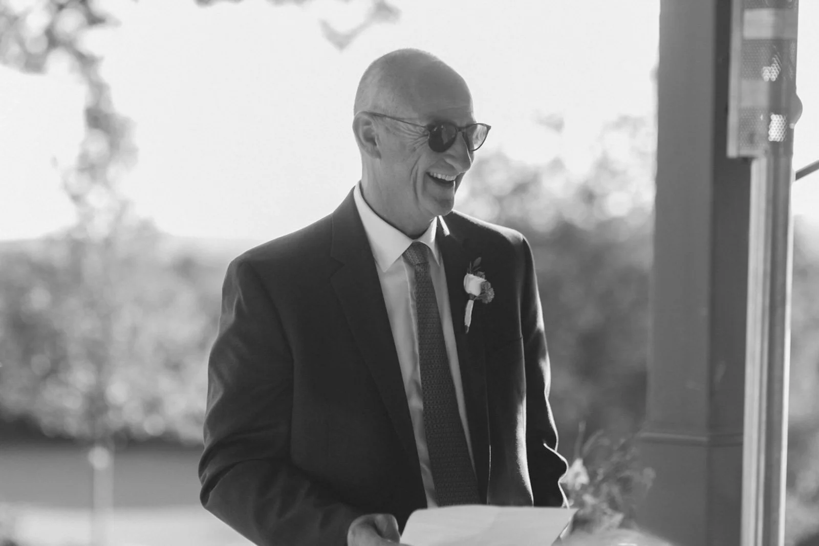 A middle-aged man in a suit, wearing sunglasses and a boutonniere, smiling while holding a piece of paper, standing indoors with sunlight and outdoor scenery in the background.