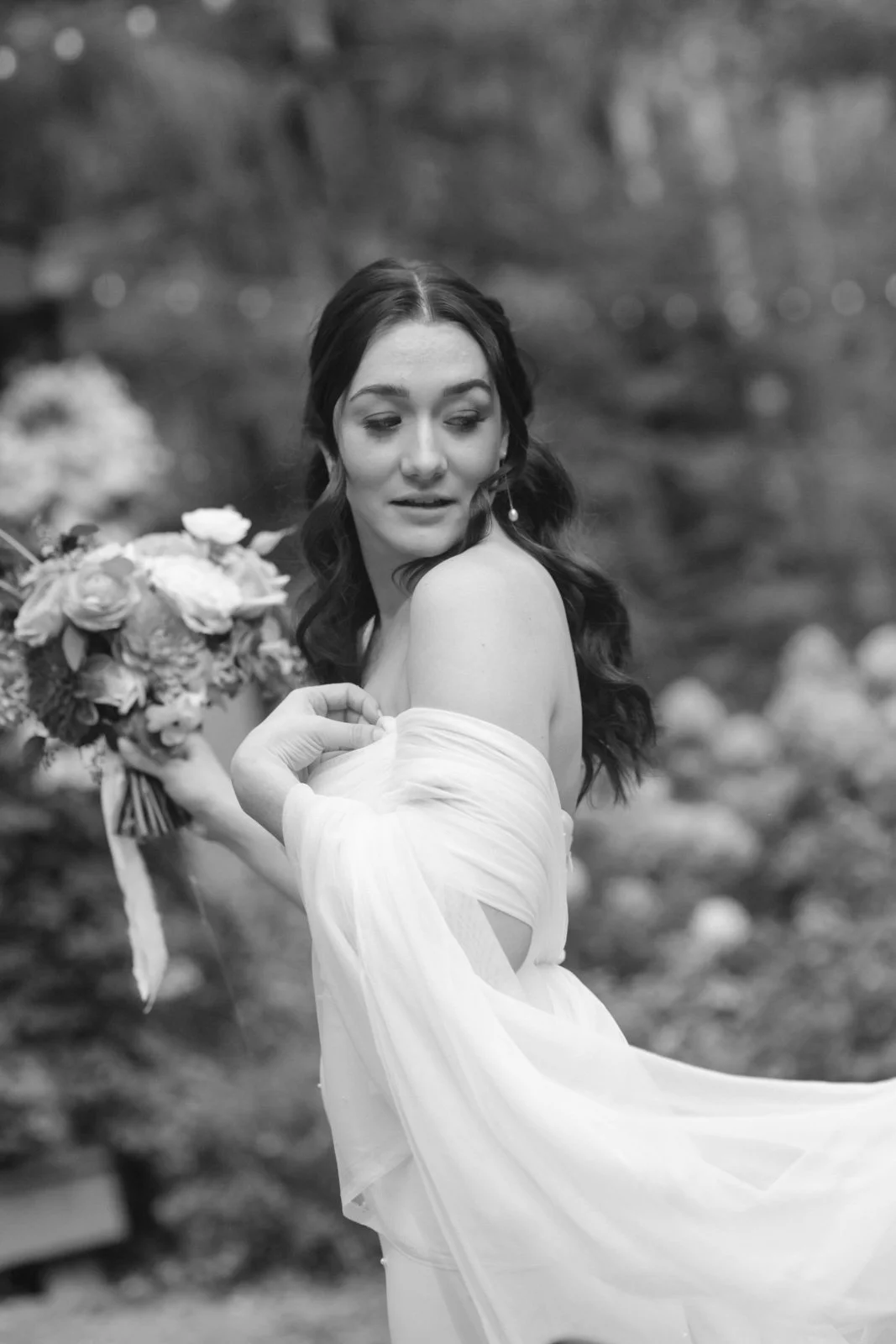 A black-and-white photo of a woman in a wedding dress holding a bouquet of flowers, looking over her shoulder outdoors.