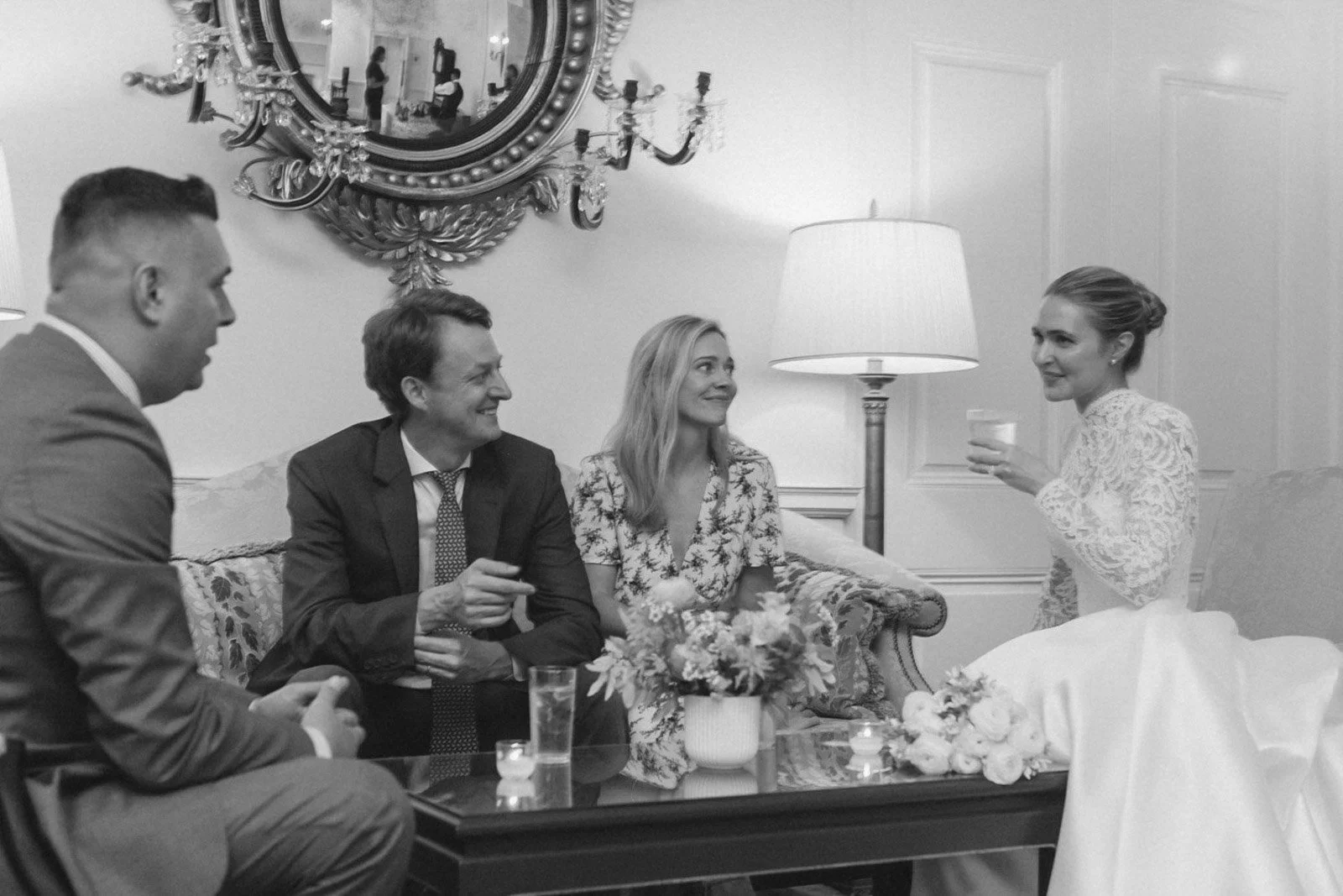 Black and white photo of four people in a living room. Three are seated on a sofa, smiling and engaging, while a woman standing on the right holds a drink and talks. A large mirror with ornate frame and chandelier are visible in the background, with 
