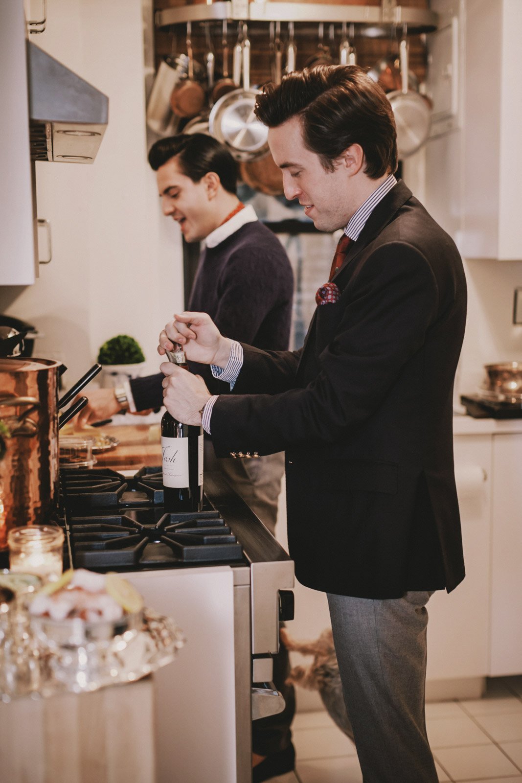 Two men preparing wine in a kitchen, one opening a bottle and the other smiling, with hanging pots and pans in the background.