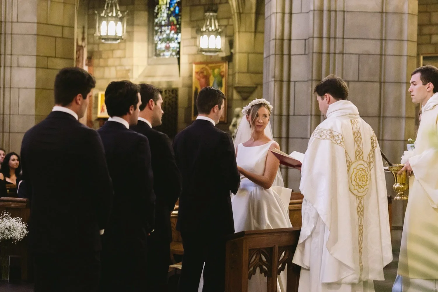 A wedding ceremony taking place inside a church with a bride, groom, and six groomsmen standing in front of a priest who is conducting the service.