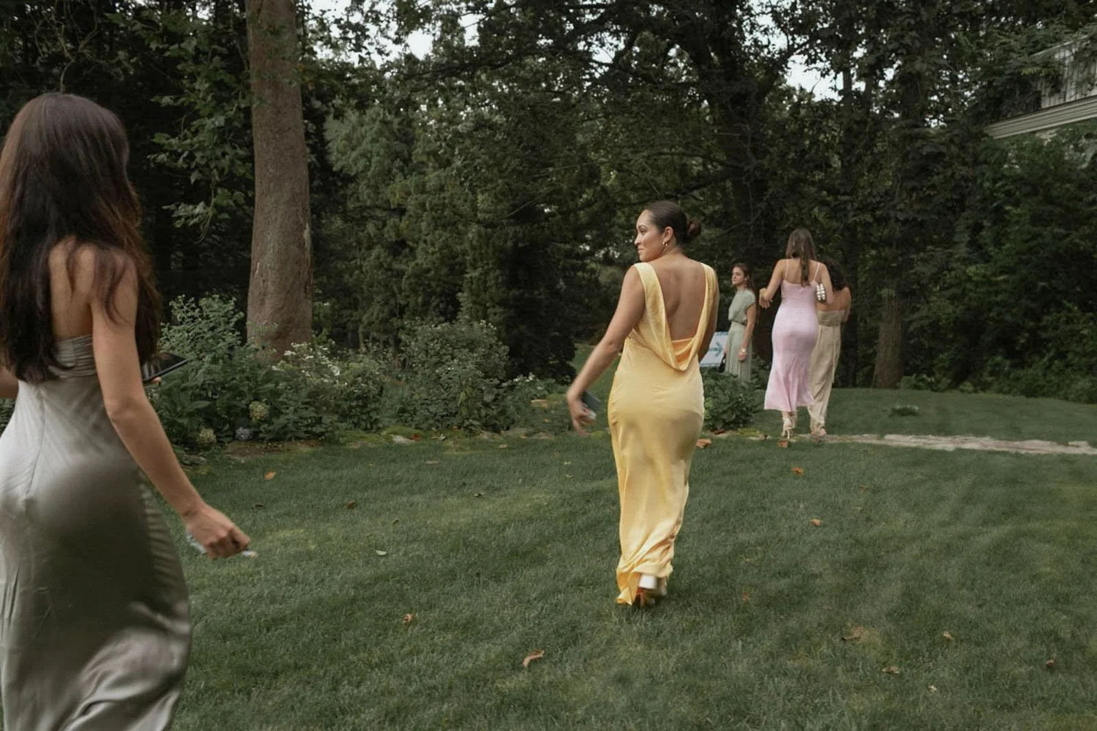 A woman in a yellow gown walking across a grassy yard with trees and bushes in the background, while other women in formal dresses are present nearby.