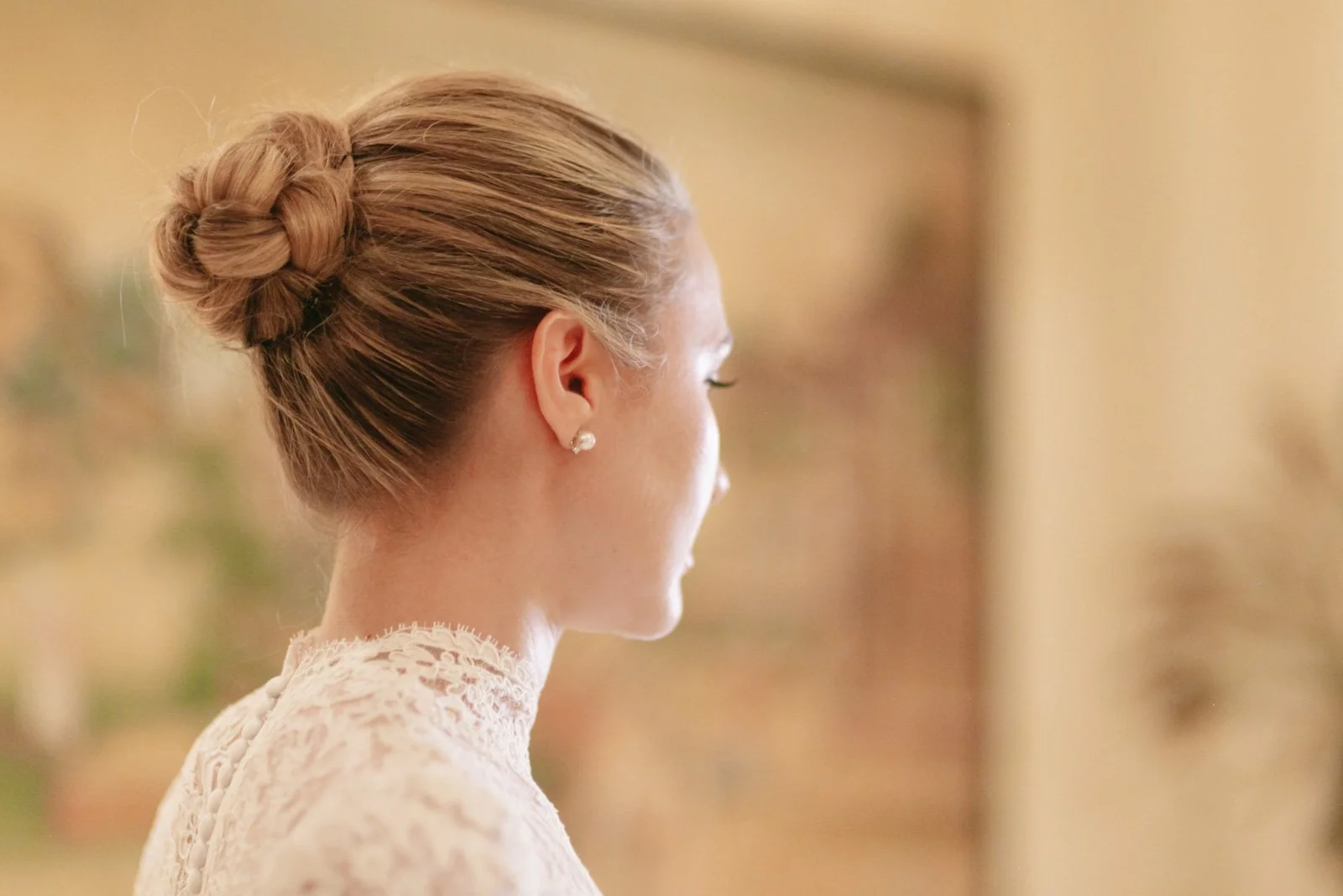 Side profile of a woman with a braided updo hairstyle, wearing pearl earrings and a white lace dress, indoors with a blurred background.