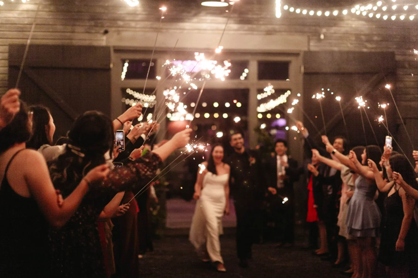Couple walking through a crowd holding sparklers at night during a celebration or wedding reception.