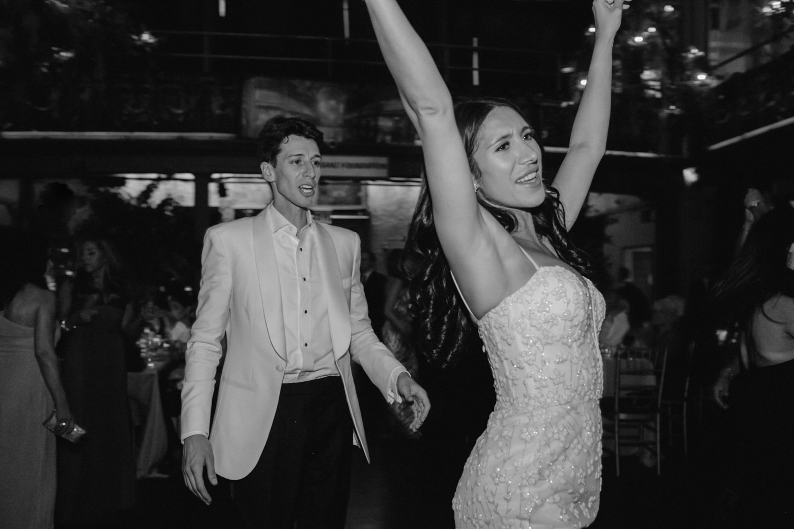 A woman in a wedding dress with her arms raised, and a man in a white tuxedo jacket in the background, at a wedding reception.