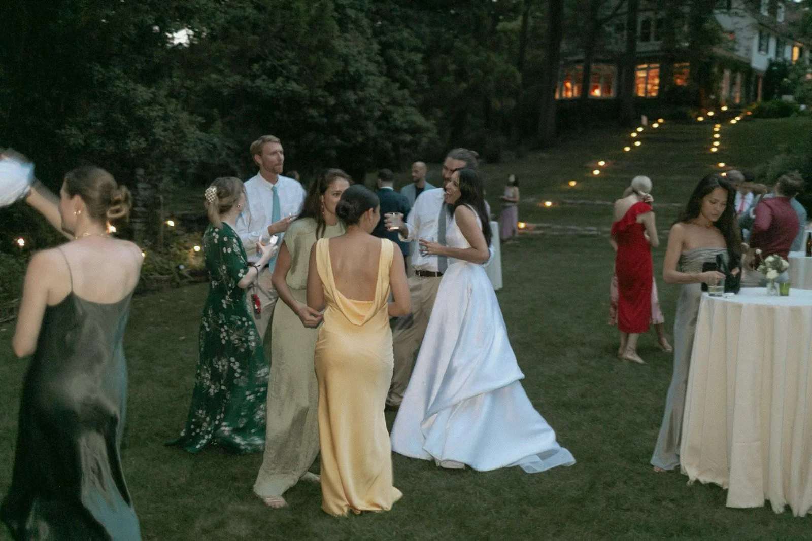 People dressed in formal attire at an outdoor evening gathering on a grassy area, with some people seen near a table with drinks and flowers, and a house with lit windows in the background.
