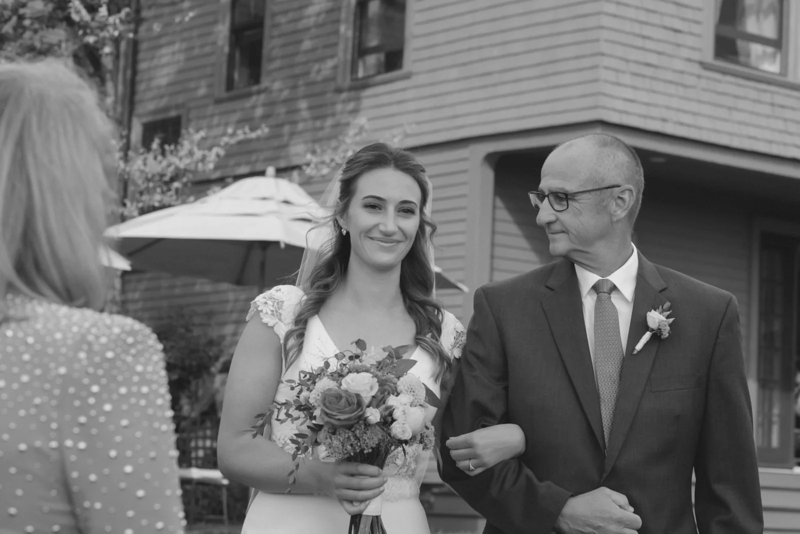A bride holding a bouquet and walking arm-in-arm with an older man while talking to a woman with back to camera, in an outdoor setting with a house and umbrella in the background.