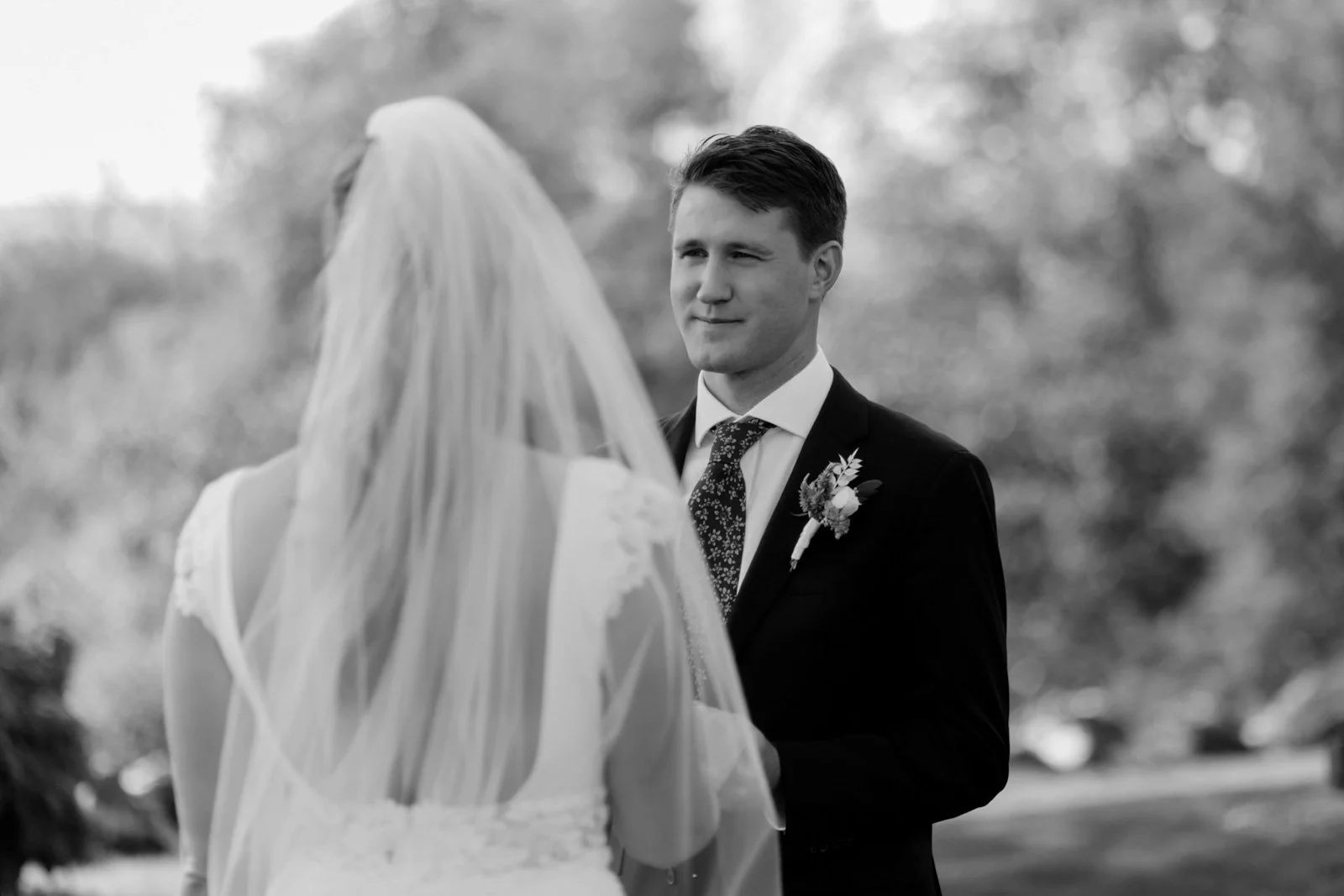 A groom in a suit with a boutonniere looking at a bride in a wedding dress and veil outdoors in black and white.