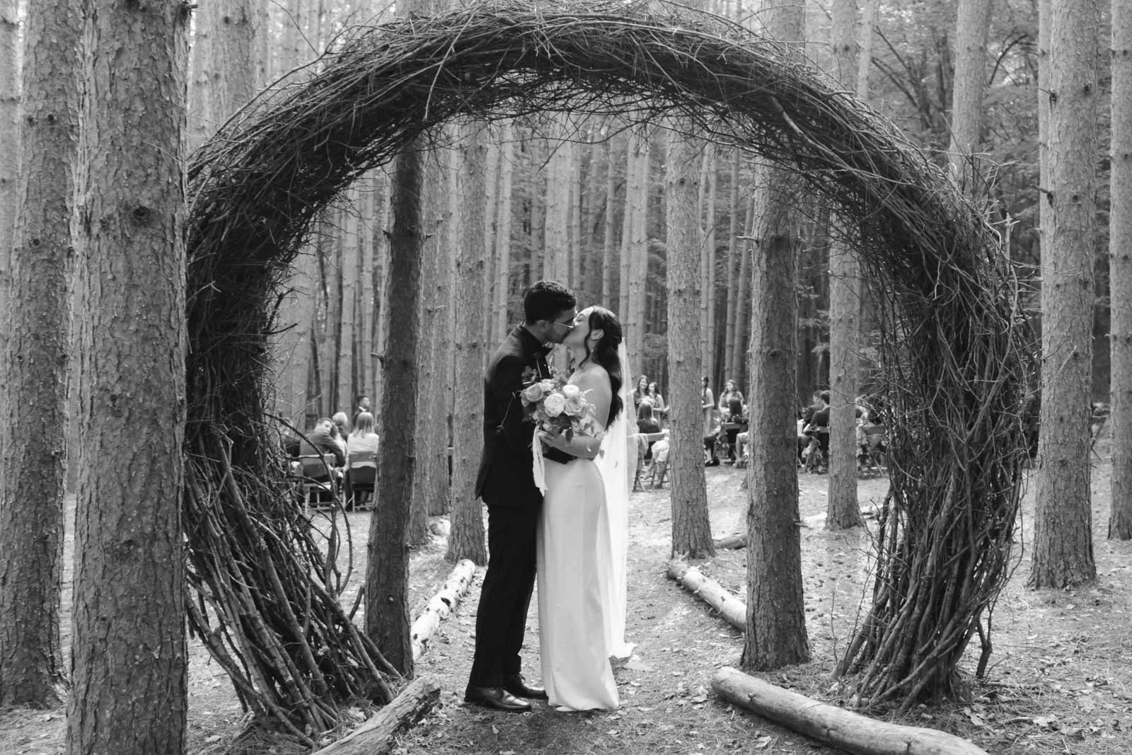 A black and white photo of a couple kissing during their outdoor wedding in a wooded area, framed by a large circular twig arrangement, with guests seated in the background.