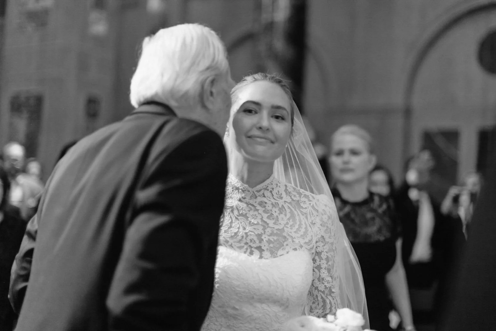A bride in a lace wedding dress and veil smiling as she looks at an older man during a wedding ceremony in a church.