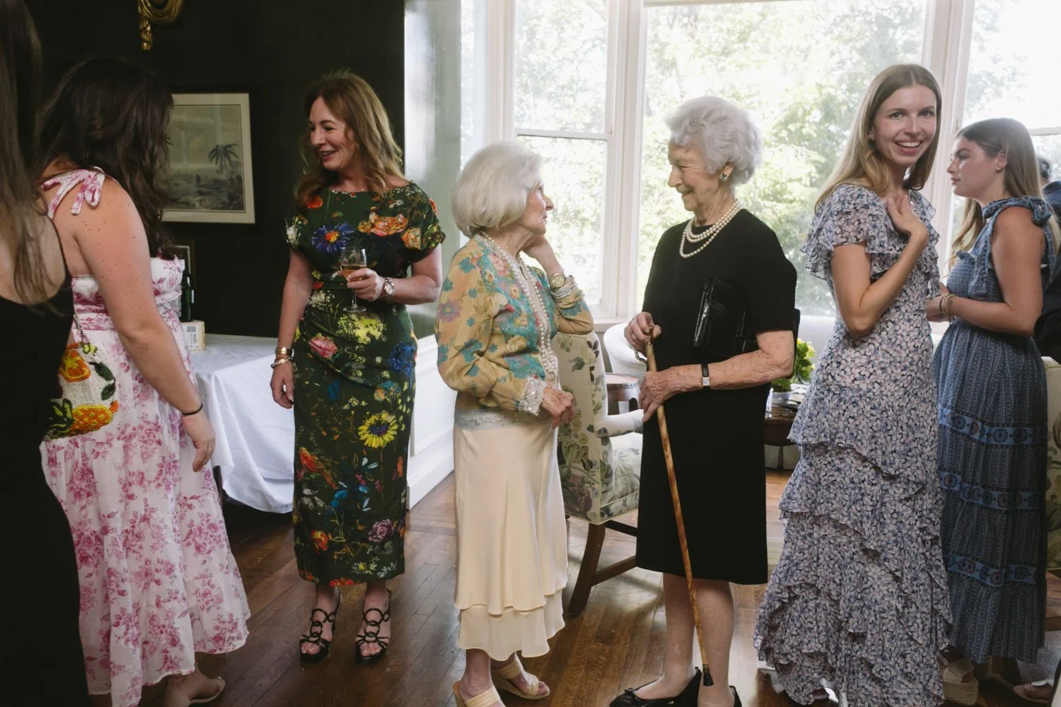 Group of women, including senior women, socializing in a well-lit room at a wedding rehearsal dinner. They are smiling, chatting, and holding drinks, with large windows showing greenery outside.