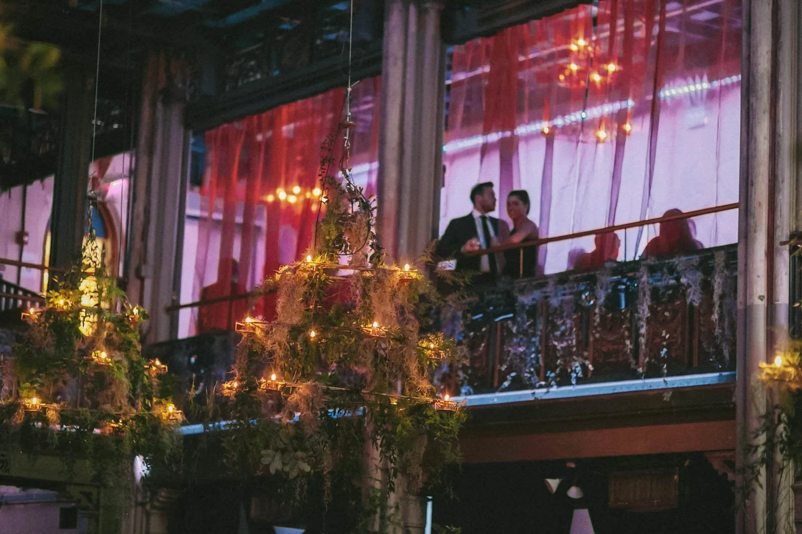 A couple in formal attire stands on an upper balcony, with a view of chandeliers and pink drapery in the background. The foreground features hanging lights and greenery, creating an elegant, festive atmosphere.